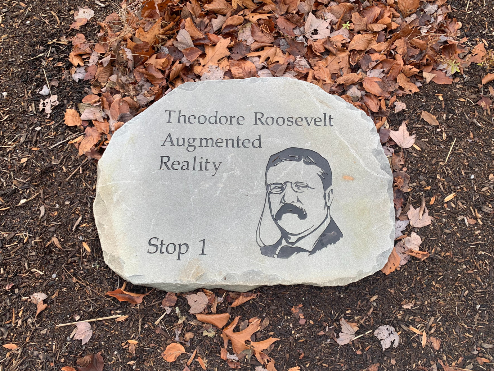 A large, flat stone on the ground with a bust of Theodore Roosevelt engraved into it. Engraved text on the stone reads: Stop 1