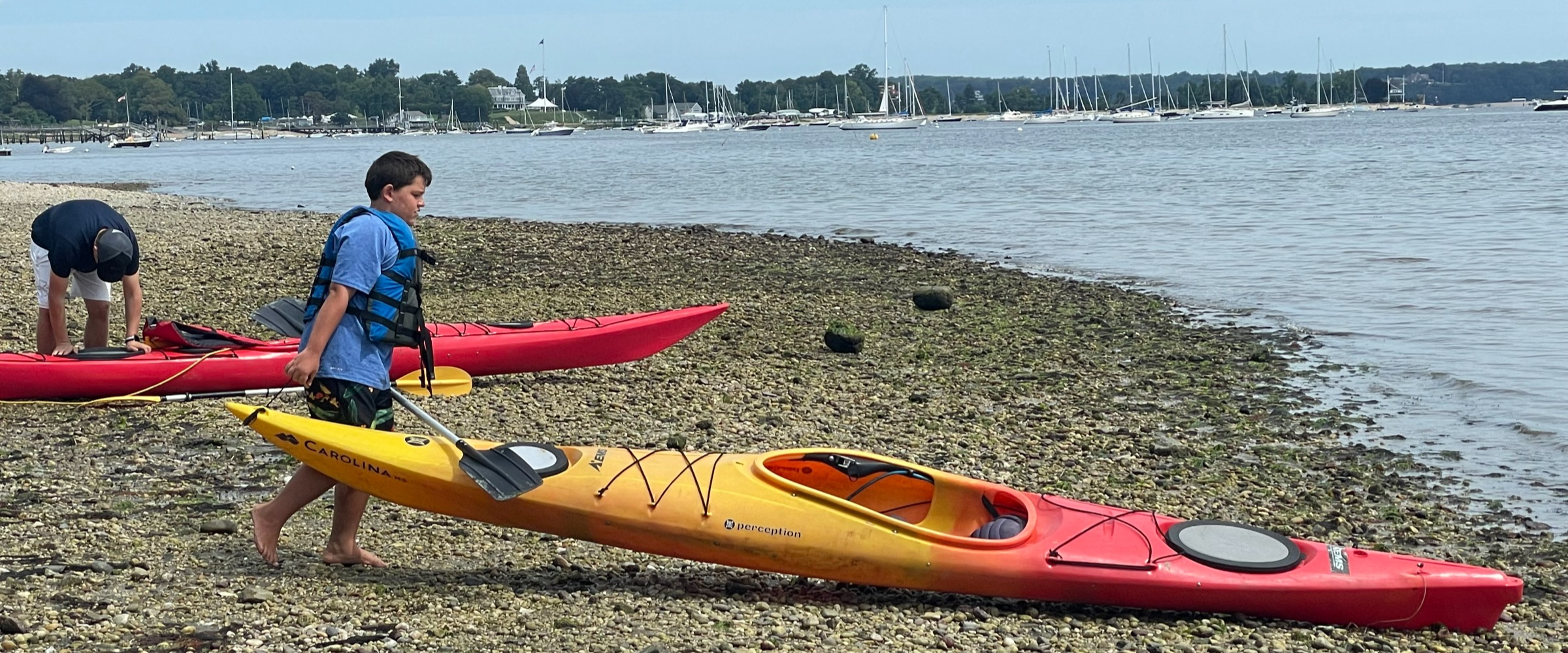 At the edge of a rocky shoreline, a child pushes a yellow kayak into the water.