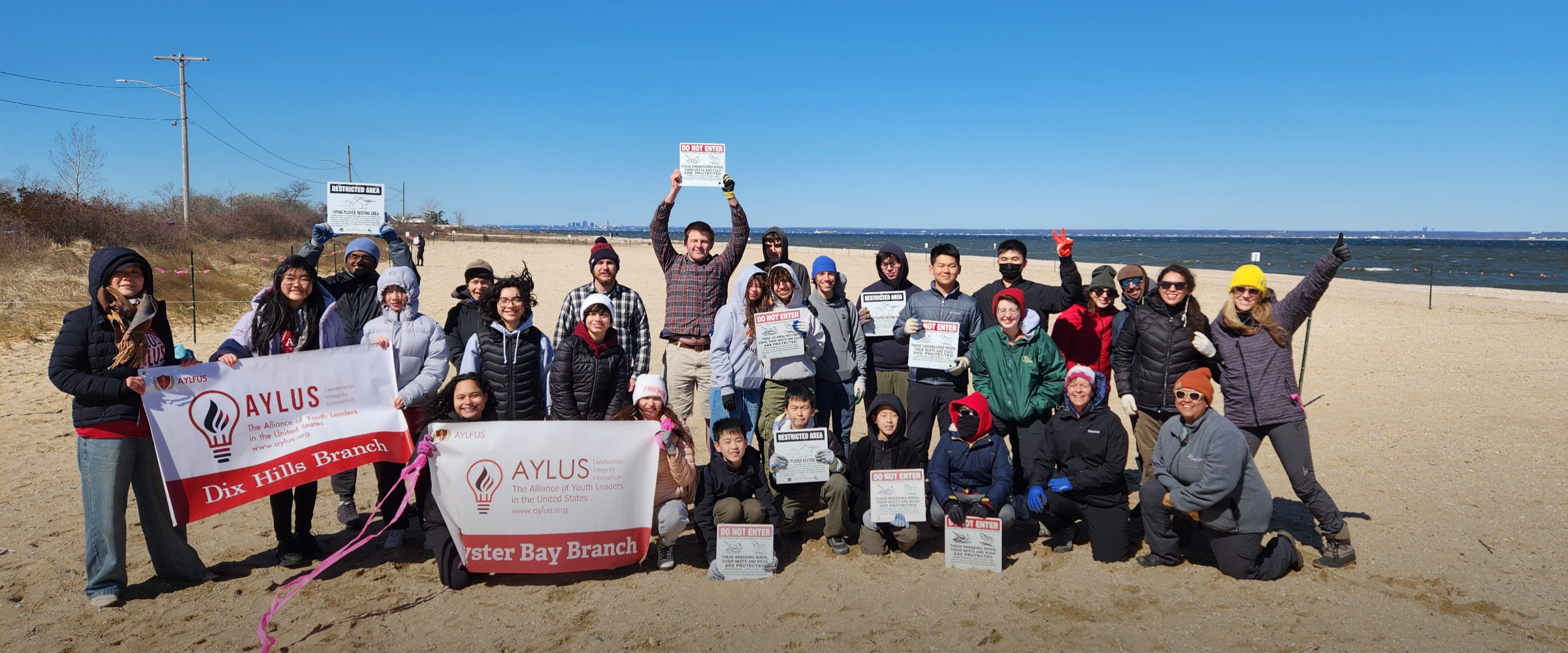 A large group of people stand together on a beach, dressed for cold weather. They are smiling.