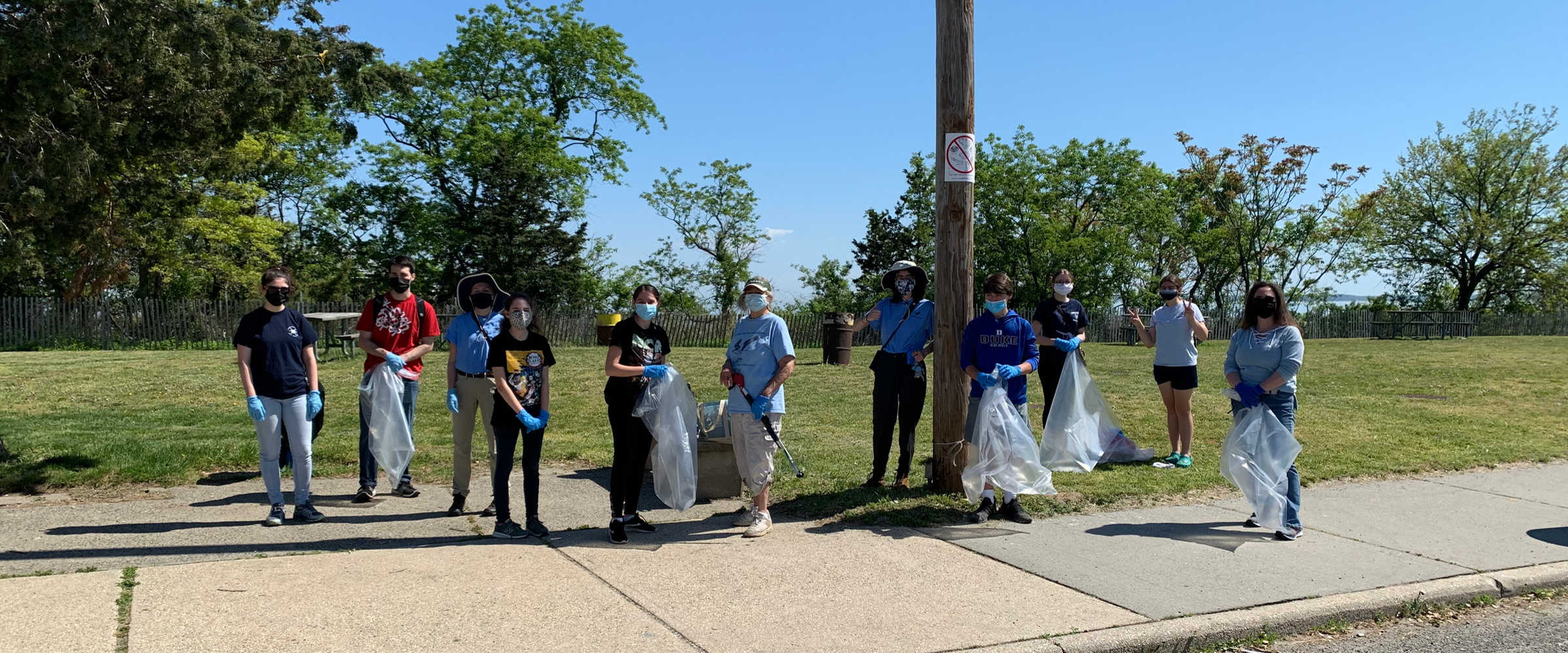 A group of people stand spread out along a sidewalk in front of a grassy park area. They are holding tools and plastic bags before setting off on a beach cleanup.